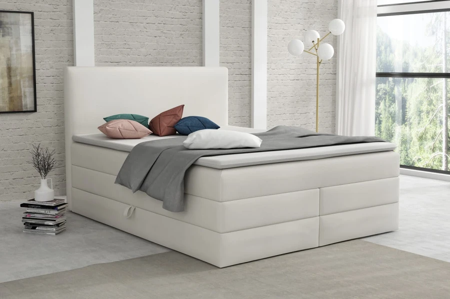 Elegantes weißes Boxspringbett mit weichem Stoffbezug vor einer hellen Backsteinwand im Schlafzimmer.
