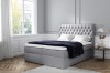 Graues Boxspringbett mit gestepptem Kopfteil und weißer Bettwäsche in hellem Raum.
