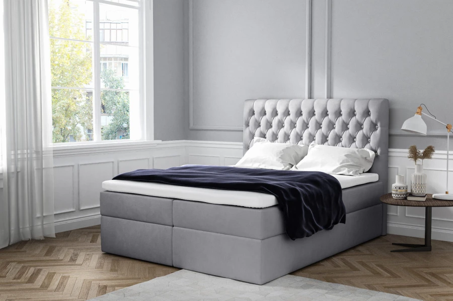 Graues Boxspringbett mit gestepptem Kopfteil und weißer Bettwäsche in hellem Raum.