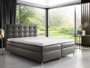 Graues Boxspringbett mit hohem Kopfteil und weißer Matratze in hellem Zimmer mit Ausblick.