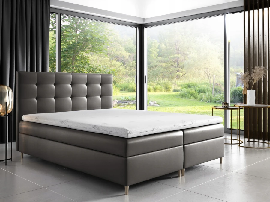 Graues Boxspringbett mit hohem Kopfteil und weißer Matratze in hellem Zimmer mit Ausblick.