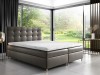 Boxspringbett in Taupe mit quadratischer Steppung vor bodentiefen Fenstern in der Natur.