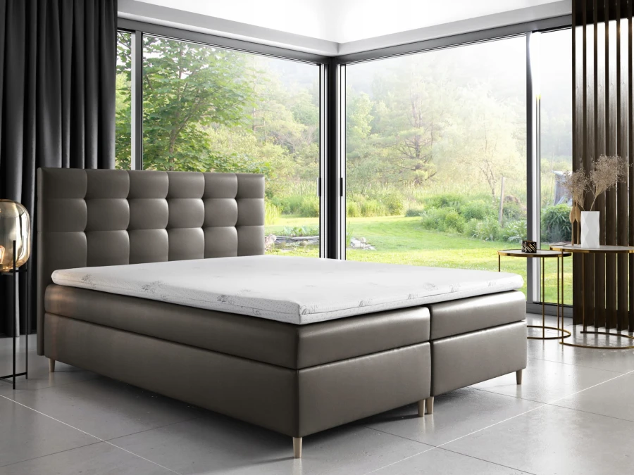 Boxspringbett in Taupe mit quadratischer Steppung vor bodentiefen Fenstern in der Natur.