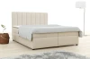 Beiges Boxspringbett mit hohem Kopfteil in hellem Schlafzimmer mit Teppich und Lampe.
