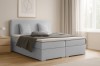Gemütliches Boxspringbett in Hellgrau mit Kissen und Decke in einem hellen Schlafzimmer.