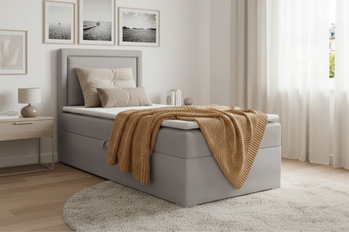 Hellgraues Boxspringbett mit Bettkasten, Topper und senfgelber Decke vor weißer Wand.